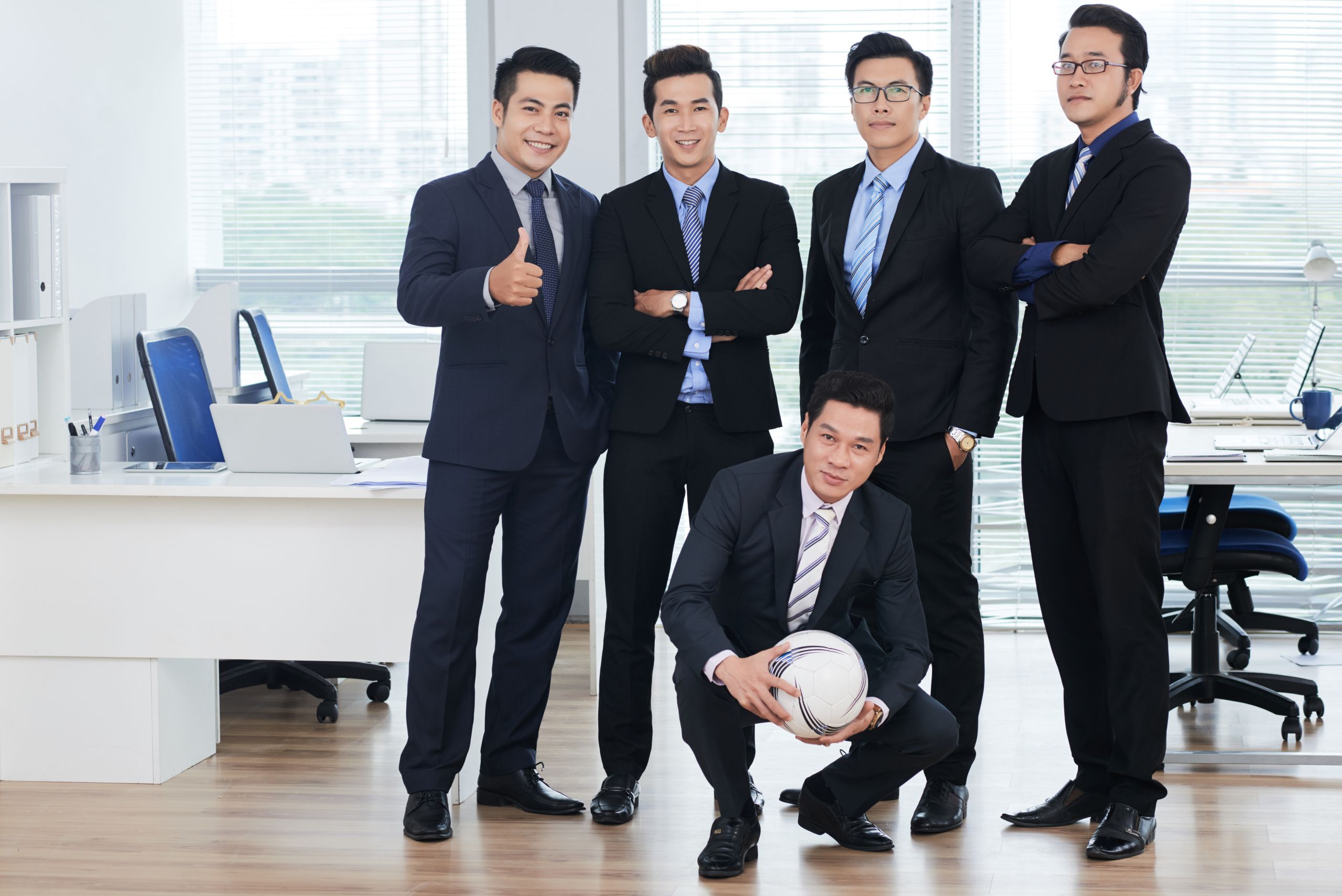 Smiling Asian managers wearing classical suits posing for photography while standing at open plan office, one of them crouching and holding soccer ball in hands