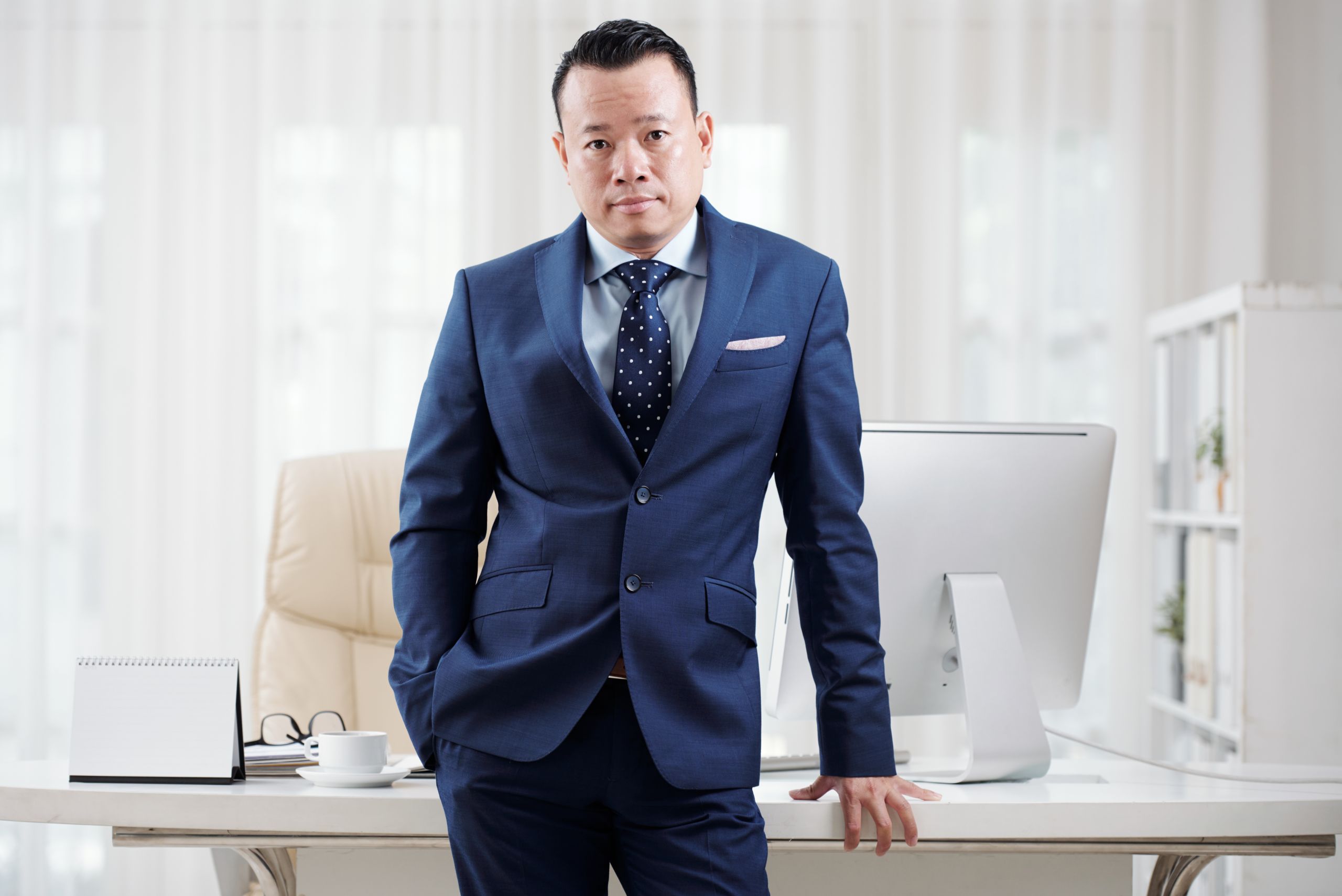 Portrait of confident Vietnamese businessman in suit leaning on his table and looking at camera
